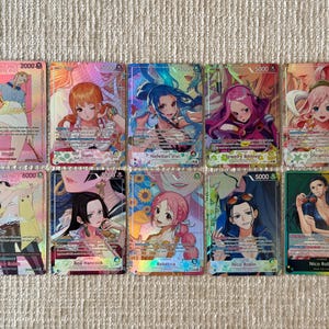 May include: A collection of ten One Piece trading cards arranged in two rows. Each card features a different female character from the series, including Nami, Nico Robin, and Boa Hancock, with colorful anime-style illustrations and text.