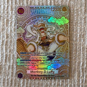 Puede incluir: Una carta coleccionable holográfica de One Piece que muestra a Monkey D. Luffy en su forma Gear 5. La carta muestra un nivel de poder de 12000, un coste de 10 y texto detallado que describe las habilidades de juego. El fondo tiene patrones dorados y azules.