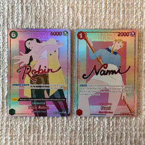 May include: Two One Piece Heroines trading cards featuring Nico Robin and Nami. The cards have a holographic finish, displaying character illustrations, names in cursive script, power levels, and game text. The cards are rectangular with rounded corners.