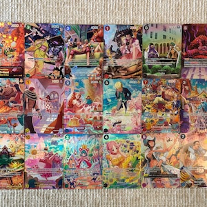 May include: A collection of fifteen One Piece trading cards arranged in a grid, each featuring colourful anime-style illustrations of various characters, numerical power stats, and text in Japanese and English, showcasing diverse scenes and settings.