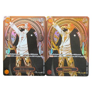 Peut inclure: Deux cartes à collectionner avec le personnage Shanks. Les cartes ont un fond doré et marron avec une illustration du personnage. Le texte sur la carte comprend "10", "12000" et "Shanks". Les cartes sont de l'ensemble "The Four Emperors/Rebellious Pirates".
