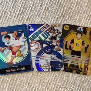 Puede incluir: Tres cartas coleccionables de One Piece que muestran a Monkey D. Luffy con varios atuendos deportivos. Las cartas presentan el logotipo de los LA Dodgers, imágenes de béisbol y la marca BVB 09. Cada carta incluye ilustraciones, texto y estadísticas.