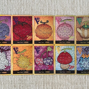 May include: A collection of ten rectangular trading cards featuring colorful illustrations of various stylized fruits with swirling patterns. Each card displays a unique fruit design, a Japanese character in the top left corner, and the text Your Turn +1000.