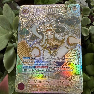 Op de afbeelding: Een holografische ruilkaart met Monkey D. Luffy van The Four Emperors/Straw Hat Crew. De kaart heeft een goud- en zilverontwerp met Japanse tekst en een karakterillustratie. De waarde van de kaart is 2000.