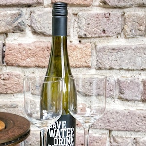 Könnte beinhalten: Zwei klare Weingläser und eine Flasche Riesling-Wein auf einem Satz von zwei rostigen Metalluntersetzern. Das Flaschenetikett lautet "Save Water Drink Riesling".