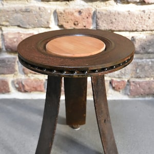 Könnte beinhalten: Ein rustikaler, dreibbeiniger Hocker aus einem wiederverwendeten Bremsscheibenrohling mit einer Holzplatte. Der Hocker hat eine dunkelbraune, rostige Oberfläche und steht auf einem grauen Betonboden.