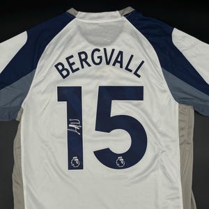 May include: White and navy blue soccer jersey with the name "BERGVALL" and the number "15" printed on the back. The jersey has a signature on the number. The sleeves are navy blue and gray.