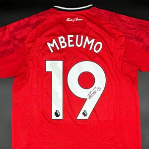 May include: Red soccer jersey with the name "MBEUMO" and the number "19" in white. The jersey has a signature below the number and the words "Theatre of Dreams" at the top.