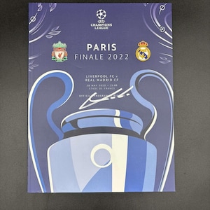 May include: A blue program booklet for the UEFA Champions League Final 2022 in Paris, France. The cover features the UEFA Champions League logo, the team logos for Liverpool FC and Real Madrid CF, and the text "Paris Finale 2022".