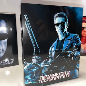 May include: Movie box set featuring the film "Terminator 2: Judgment Day." The cover art shows a man in sunglasses holding a shotgun on a motorcycle. The title is in white and red text. Other movie boxes are visible.