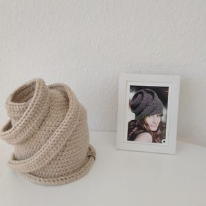 Könnte beinhalten: Ein beigefarbener gehäkelter Cowl-Schal liegt auf einer weißen Oberfläche. Der Schal ist aus dickem Garn gefertigt und hat ein gerolltes Design. Rechts befindet sich ein gerahmtes Foto einer Frau mit ähnlichem Hut.