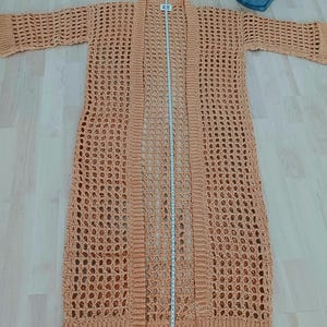 Könnte beinhalten: Ein langer, hellbrauner Häkelcardigan mit lockerer Passform und offenem Vorderteil. Der Cardigan ist aus einem zarten, durchbrochenen Stoff gefertigt und hat einen lässigen, bohemian Stil.