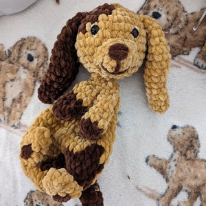 May include: A handmade crochet dog plushie in shades of brown and tan. The dog has floppy ears, a small nose, and black eyes. The plushie is lying on a white blanket with a dog pattern.