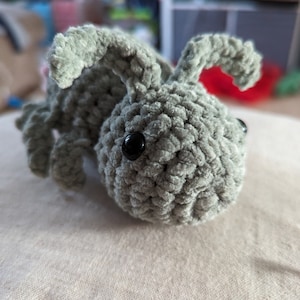 May include: A gray crocheted bunny with black eyes. The bunny is lying on a white surface.