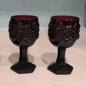 Peut inclure: Deux verres à pied en verre bordeaux profond avec un motif géométrique en relief sur le calice. Chaque verre possède une tige épaisse et une base hexagonale. Le verre est sombre et translucide, affichant un motif répétitif de cercles.