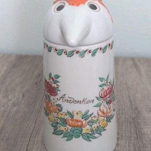 May include: A white ceramic pitcher with a lid shaped like a bird head featuring a red crest and black eyes. The body is decorated with a floral wreath pattern in green, orange, and pink, and the word Andenken is written in cursive script.