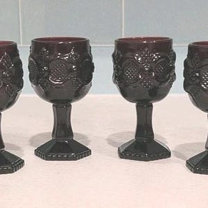 May include: A set of four deep burgundy wine glasses made of pressed glass. Each glass features a raised, textured floral and geometric pattern around the bowl, a smooth stem, and a hexagonal base with a beaded edge detail at the bottom.