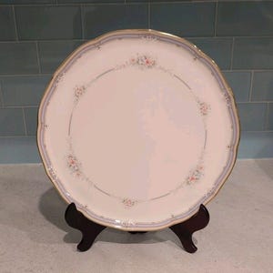 May include: A round porcelain dinner plate with a scalloped edge and a gold-trimmed rim. The plate features a white center decorated with a delicate floral garland of pink and blue flowers, accented by a soft blue border line around the perimeter.