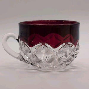 Puede incluir: Una taza de cristal con una base transparente con un patrón de corte de diamante y una banda de color rojo intenso alrededor del borde superior. La taza incluye un asa curva en el lado izquierdo, con el cristal transparente hacia el borde rojo.