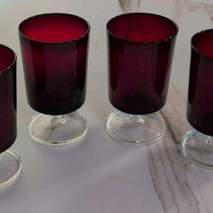 May include: A set of four matching drinking glasses, each featuring a deep ruby red cylindrical bowl supported by a clear glass stem and a circular base. The glasses are arranged in a row, showcasing the consistent color and structural design.
