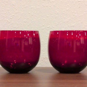 May include: Two rounded, deep red glass candle holders with a smooth, glossy finish. Each vessel features a wide, open top and a curved, bulbous body that tapers toward the base, reflecting light across the vibrant, translucent crimson surface.