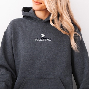 May include: Dark heather gray hoodie with a kangaroo pocket and a drawstring hood. The word "squirrel" is printed in white, along with a small squirrel graphic. The hoodie is paired with light blue jeans.