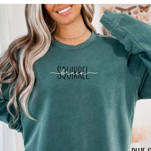 May include: A teal-colored sweatshirt with the text "Squirrel" in a cursive font above the word "birch" in a sans-serif font. The words "Blue Spruce" are printed below the sweatshirt. The sweatshirt has long sleeves and a crew neck.