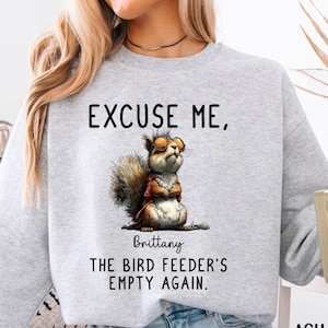 May include: Light gray sweatshirt with the text "EXCUSE ME, THE BIRD FEEDER'S EMPTY AGAIN." and a cartoon squirrel wearing glasses. The word "Brittany" is below the squirrel. The word "ASH" is in the bottom right corner.
