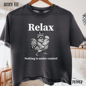 May include: Dark gray t-shirt with the word "Relax" in white. Below the word "Relax" is a graphic of a raccoon riding a rooster. The text "Nothing is under control" is below the graphic. The shirt is hanging on a wooden hanger.