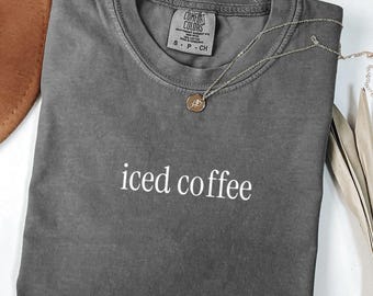 May include: A gray Comfort Colors t-shirt with the words "iced coffee" in white. The shirt has a crew neck and short sleeves. A gold necklace with a pendant rests on the shirt. The word "PEPPER" is at the bottom left.