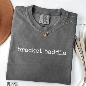 May include: A gray t-shirt with the text "bracket baddie" in white. A silver necklace with a pendant is draped across the shirt. The shirt is labeled "Pepper" at the bottom.