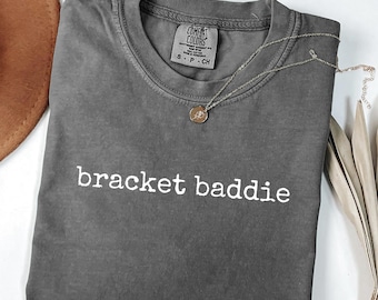 Może przedstawiać: Szara koszulka z białym napisem "bracket baddie". Srebrny naszyjnik z wisiorkiem jest zarzucony na koszulkę. Koszulka jest oznaczona jako "Pepper" na dole.