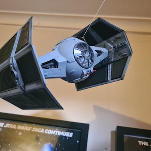 Puede incluir: Maqueta de un caza TIE de Star Wars, suspendida de un hilo. La nave espacial es gris con paneles solares negros y una cabina detallada. La imagen incluye un póster enmarcado con el texto "THE STAR WARS SAGA CONTINUES."