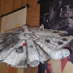 May include: A detailed model of the Millennium Falcon starship, a prominent element from the Star Wars franchise. The model is predominantly gray with red and white accents, suspended by strings. The background includes Star Wars-themed artwork.