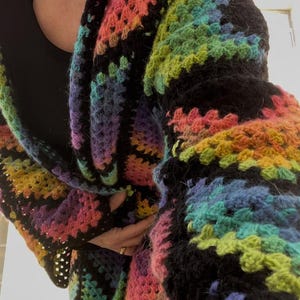 May include: A colorful, handmade crochet cardigan in a rainbow of hues, including pink, orange, yellow, green, blue, and purple. The cardigan has a black border and a textured, open weave design. A cozy and vibrant piece of wearable art.