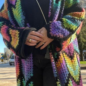 May include: A long, open-front crochet cardigan in a vibrant rainbow pattern. The colorful design features squares of pink, yellow, green, and purple, outlined in black. The cardigan is worn over a black turtleneck and black pants.