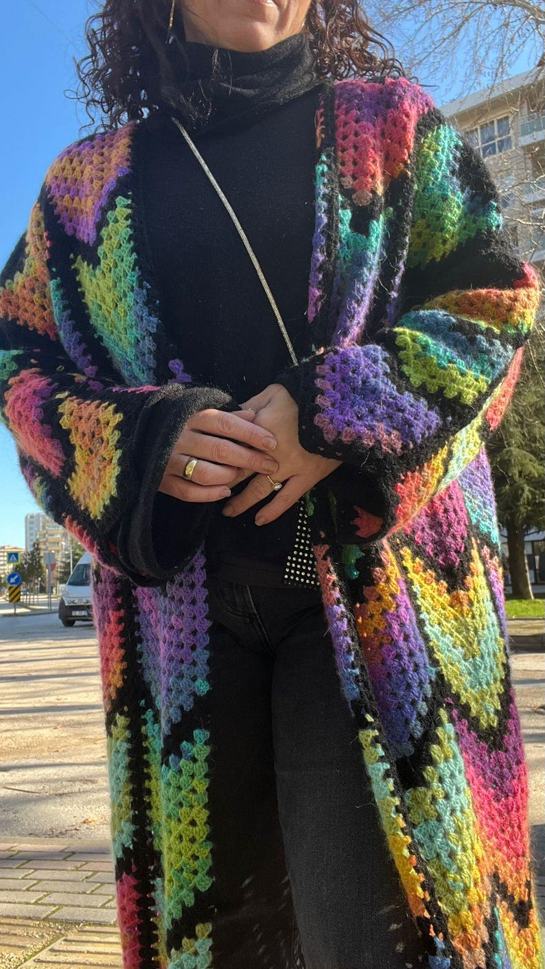 May include: A long, open-front crochet cardigan in a vibrant rainbow pattern. The colorful design features squares of pink, yellow, green, and purple, outlined in black. The cardigan is worn over a black turtleneck and black pants.
