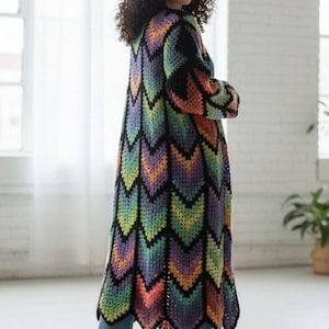 May include: A long, colorful crochet cardigan with a chevron pattern in shades of black, purple, green, yellow, and orange. The cardigan is worn over blue jeans and white sneakers. The garment has long sleeves and a scalloped hem.