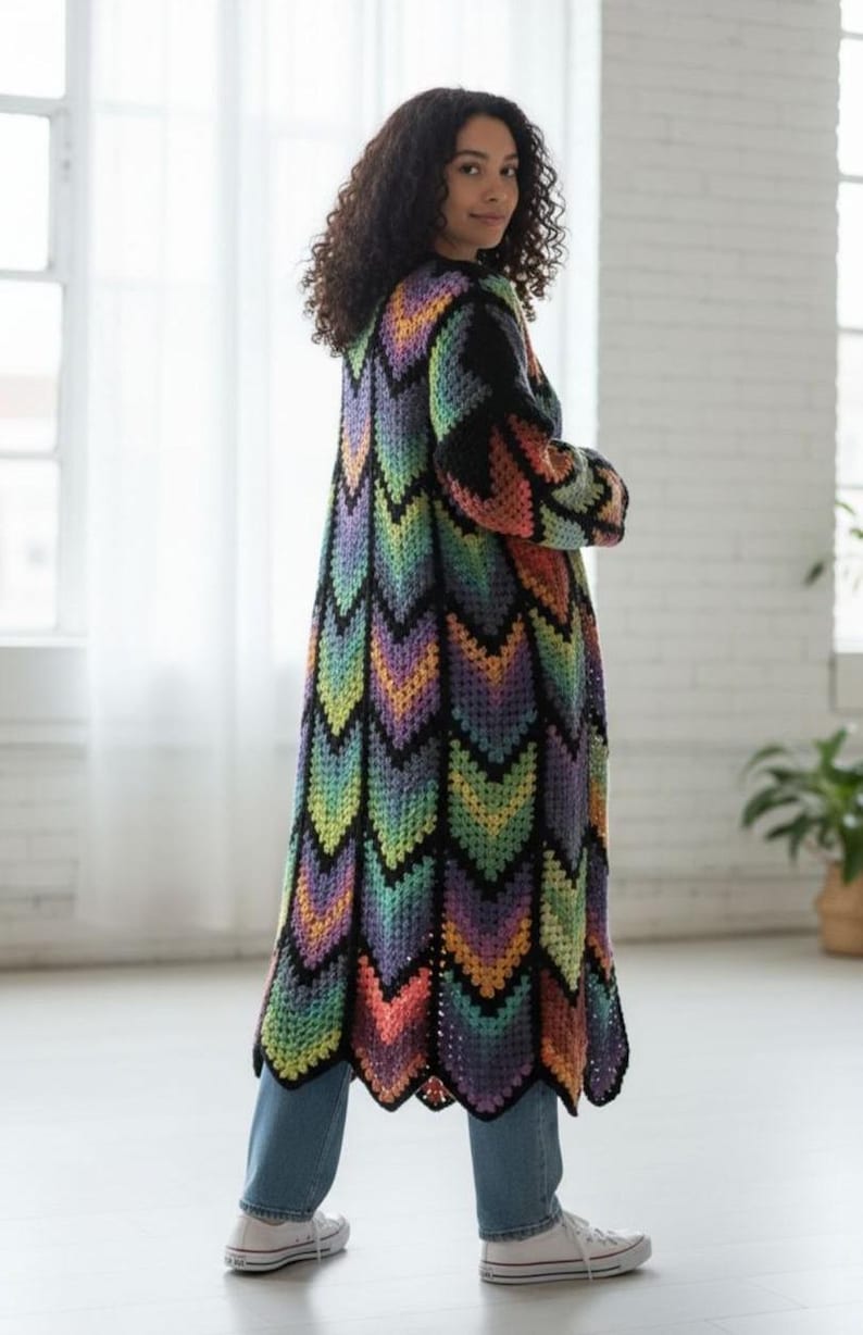 May include: A long, colorful crochet cardigan with a chevron pattern in shades of black, purple, green, yellow, and orange. The cardigan is worn over blue jeans and white sneakers. The garment has long sleeves and a scalloped hem.