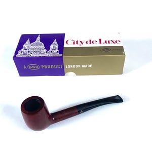 Pode incluir: Um cachimbo de fumar bordô com boquilha preta, sobre uma superfície branca. Ao lado, uma caixa com "City de Luxe" e "London Made". A caixa tem uma seção roxa com uma ilustração de igreja.