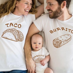 May include: A family wearing matching white t-shirts and a onesie. The shirts feature brown illustrations and text: "TACO", "BURRITO", and "TAQUITO". The illustrations depict a taco, a burrito, and a taquito.