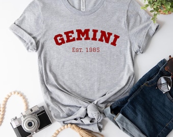 Pode incluir: Camiseta cinza com texto vermelho que diz "GEMINI EST. 1985".