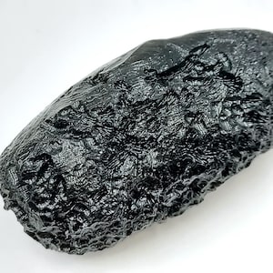 May include: A dark, glossy, black tektite with a textured surface. The elongated, oval-shaped stone has a rough, bubbly appearance, and is set against a white background. The tektite is a natural glass formed by meteorite impact.