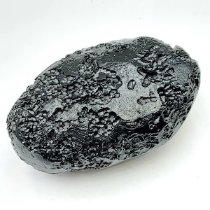 May include: A dark, oval-shaped, black stone with a textured surface. The stone has a glossy finish and a unique, organic appearance. The surface is covered in intricate patterns and ridges. This natural stone could be a decorative item or a collectible.