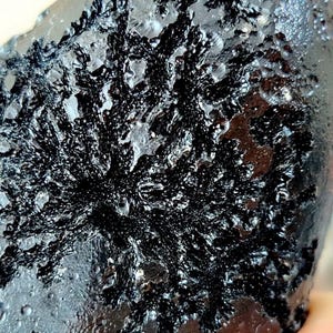 May include: Close-up of a dark rock with a textured surface. The rock has a black, intricate, branching pattern across its surface, resembling a crystallized or organic structure. The overall color is a deep black with hints of grey.