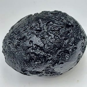 May include: A dark, irregularly shaped, black tektite with a textured surface. The natural glass object has a glossy appearance and a unique, organic form. The tektite is a natural glass formed from the impact of a meteorite.