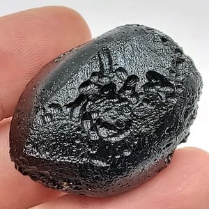 May include: A dark, irregularly shaped, black stone with a textured surface. The stone has a glossy sheen and appears to be a natural formation. The overall shape is oblong, with a slightly rounded appearance. The surface has a pitted and uneven texture.