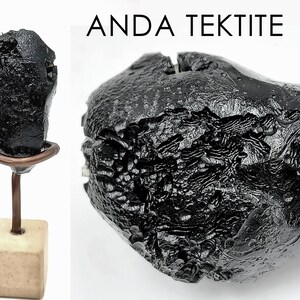 May include: A black Anda Tektite, a natural glass formed by a meteorite impact, with a textured surface. The tektite is displayed on a metal stand with a wooden base. The image includes the text "ANDA TEKTITE".