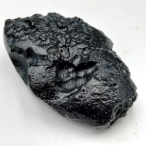 May include: A dark, irregularly shaped, black tektite with a rough, textured surface. The tektite has a glossy sheen and appears to have small, rounded protrusions and indentations across its surface. The overall shape is elongated and organic.