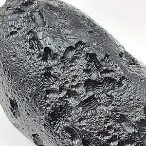 May include: Close-up of a dark, textured, oblong object. The surface is a deep black with a rough, uneven texture, featuring raised bumps and ridges. The object appears to be a natural stone or mineral.